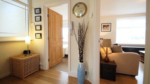 Un couloir bien éclairé avec une porte en bois, un vase bleu orné de branches, une petite table avec une lampe et un canapé également doté d’une lampe.