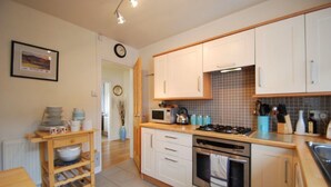 Une cuisine avec des meubles blancs, une cuisinière et un four à micro-ondes. On y trouve un chariot en bois avec des bols et des assiettes, ainsi qu’une horloge au mur.