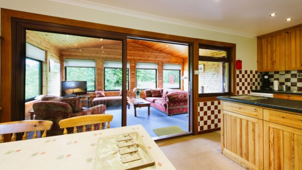 A spacious living area with a sofa and chairs, a dining table, and a kitchen with wooden cabinets and a checkered backsplash. - The Buzzard (Crianlarich)