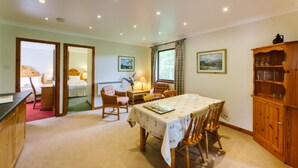 A room with a dining table, chairs, a wooden cabinet, and a bed in the adjacent room. - The Buzzard (Crianlarich)