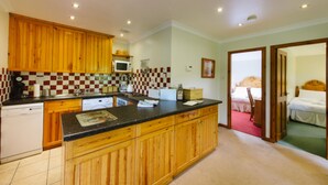 A compact kitchen with wooden cabinets, a microwave, and a sink. There is a small dining area with a table and chairs. The room has a carpeted floor and a door leading to a bedroom with a bed and a nightstand. - The Buzzard (Crianlarich)