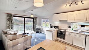 Eine moderne Küche mit weißen Schränken, eine Essecke mit Holztisch und ein Blick auf einen Garten durch große Fenster.