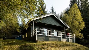 A cabin with a porch, surrounded by trees and a grassy hill. - The Osprey (Crianlarich)