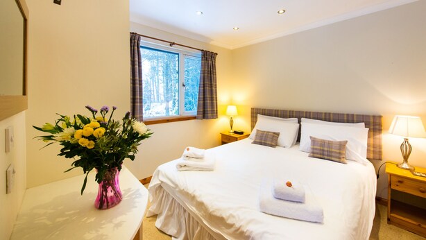 A hotel room with a bed, bedside lamps, a window with curtains, a vase with flowers, and a wall-mounted security camera. - The Osprey (Crianlarich)