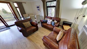 A spacious living room with leather sofas, a coffee table, and a fireplace. There are large windows providing natural light and a view of the outdoors. - The Osprey (Crianlarich)