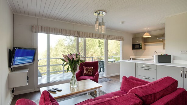 A modern living room with a red sofa, a coffee table, and a TV. There is a large window with a view of trees. - The Ptarmigan (Crianlarich)