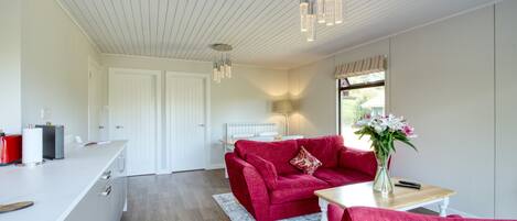 Ein modernes Wohnzimmer mit einem roten Sofa, einem weißen Couchtisch und einem Blumenstrauß. Es gibt eine Küchenzeile mit einer weißen Arbeitsplatte und einem roten Gerät. Der Raum hat Holzböden und eine weiße Decke mit Pendelleuchten.