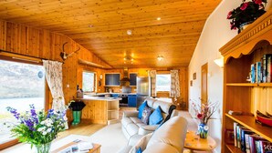 L’intérieur chaleureux d’un chalet, avec un plafond en bois, un espace de vie comprenant un canapé et une bibliothèque, une cuisine équipée d’une cuisinière et d’un évier, et une grande fenêtre offrant une vue sur l’extérieur.