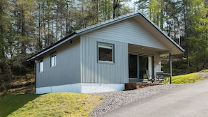 A small, single-story cabin with a sloped roof, surrounded by a forest. - The Stalker (Crianlarich)