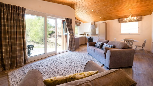 A spacious living area with a wooden ceiling, a sofa set, a dining table, and a view through large glass doors. - The Stalker (Crianlarich)