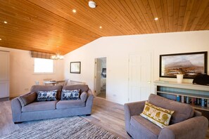 Una sala de estar con techo de madera, un sofá gris con cojines estampados, un sillón gris más pequeño, una estantería y un cuadro enmarcados con un paisaje.