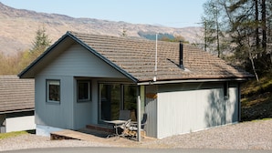 A small, single-story building with a sloped roof, a porch with a table and chairs, and a view of a mountainous landscape. - The Stalker (Crianlarich)