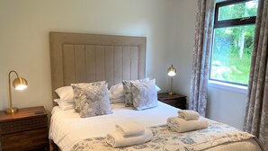 Una cama bien hecha con ropa de cama floral, dos lámparas de noche y vista a zonas verdes a través de una ventana.