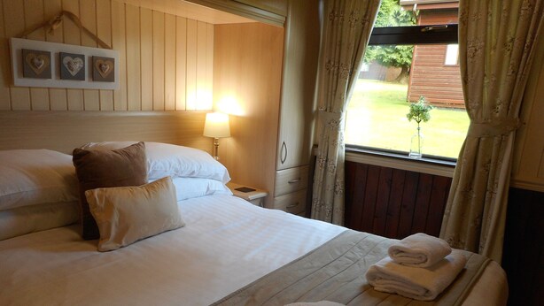 A neatly made bed with white linens and a pillow, a bedside lamp, and a window with curtains. - Birch Lodge 13 with Hot Tub (Newton Stewart)