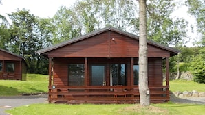 A small wooden cabin with a porch, a stone wall in the background, and a grassy area in front. - Birch Lodge 19 with Hot Tub (Newton Stewart)