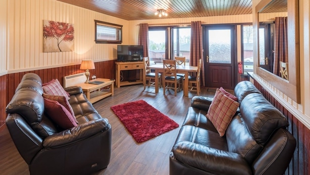 A cozy living room with leather sofas, a TV, a dining table, and a red rug. - Birch Lodge 19 with Hot Tub (Newton Stewart)