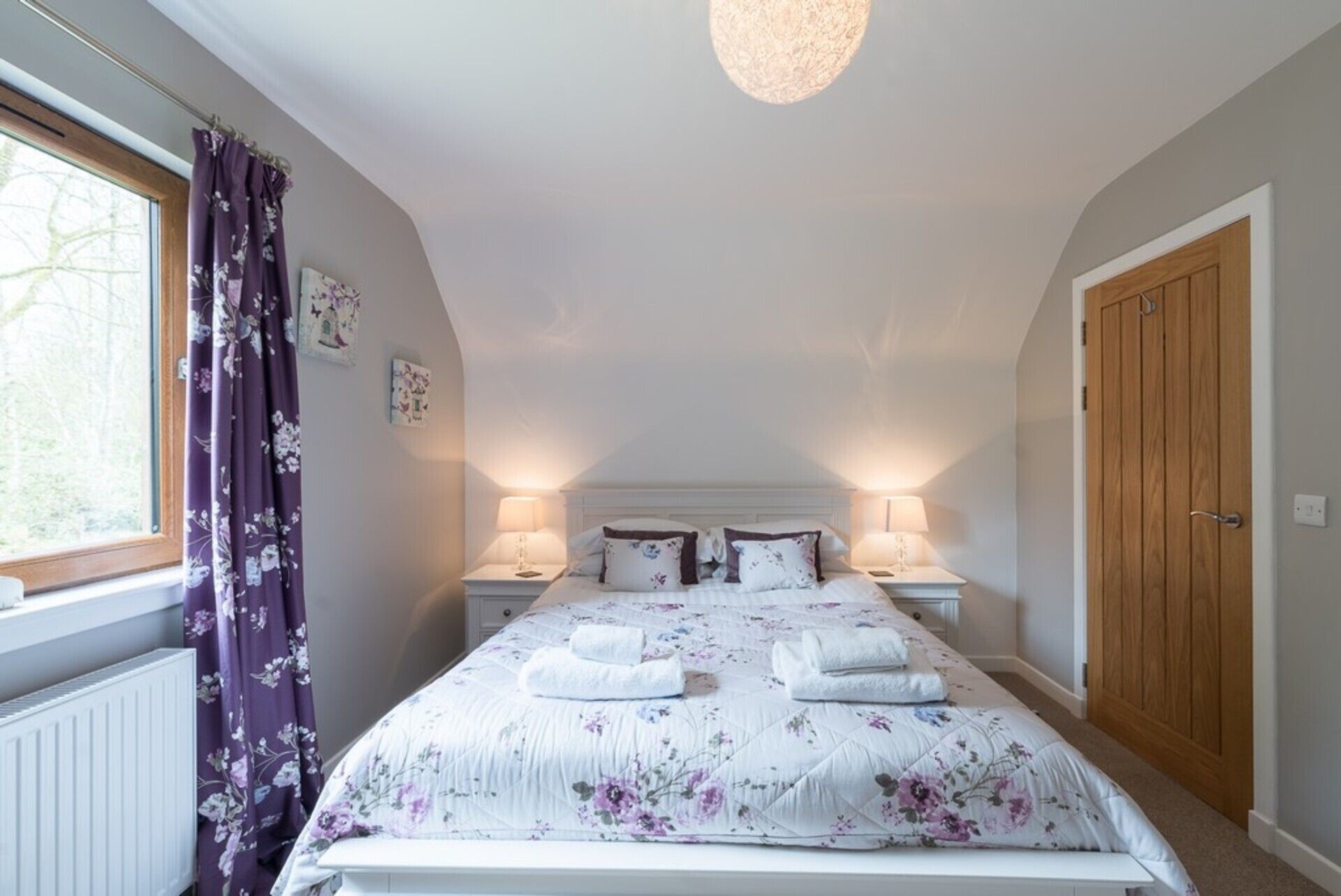 A bedroom with a large bed, two bedside tables with lamps, a wooden door, and a window with purple curtains.