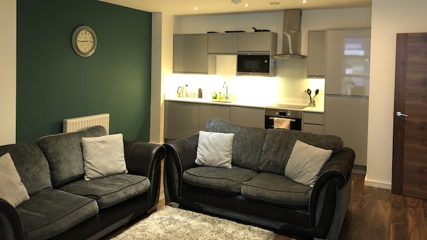 Un salon moderne avec deux canapés sombres et un tapis blanc, une cuisine avec des meubles gris et un four à micro-ondes, et une horloge au mur.
