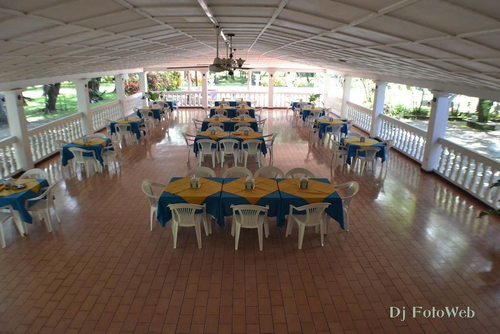 Un espacio de comedor al aire libre con techo, varias mesas y sillas, rodeado de barandas blancas y vistas a vegetación.