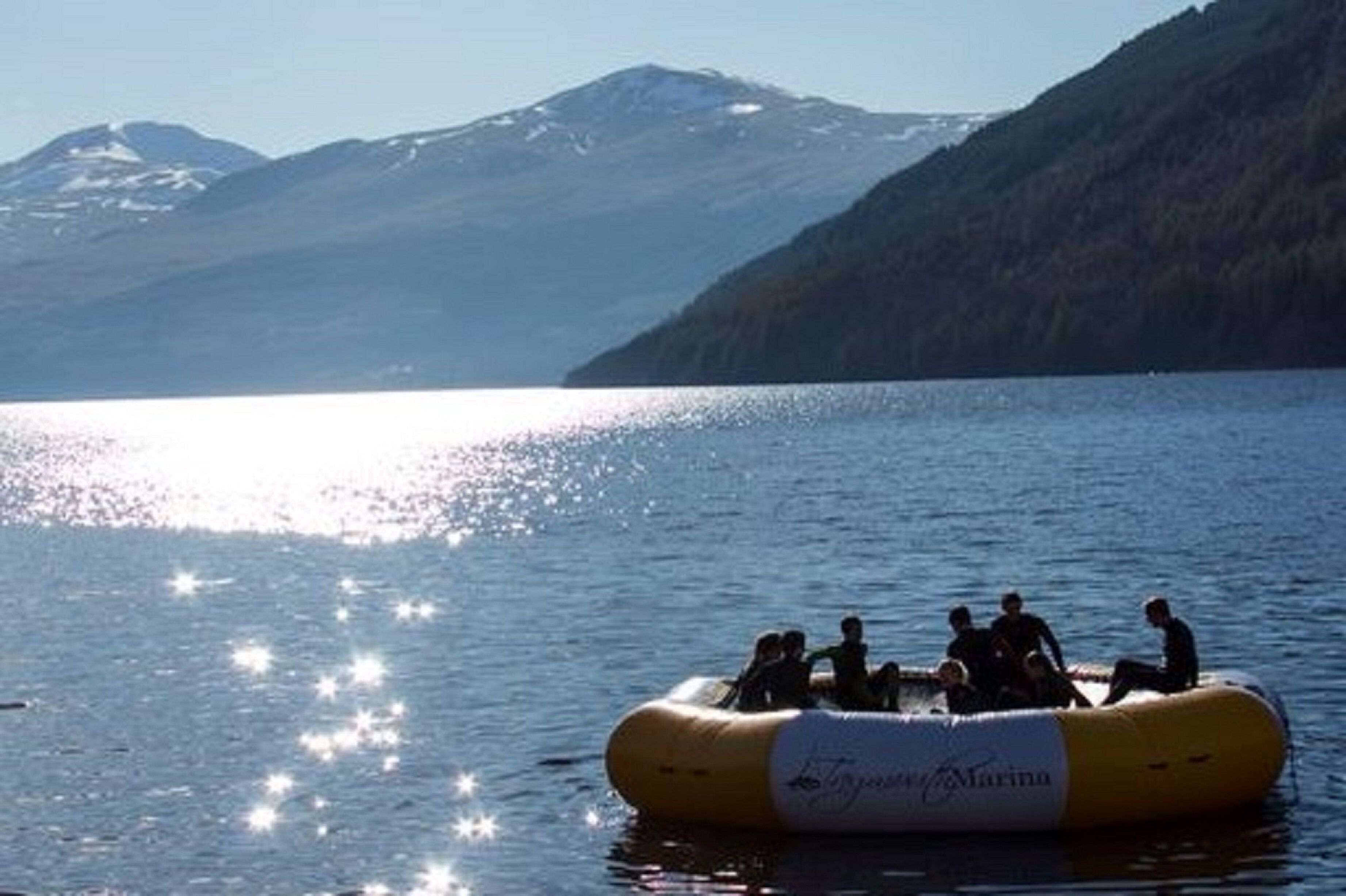 A group of people on a yellow inflatable raft on a calm lake with mountains in the background.