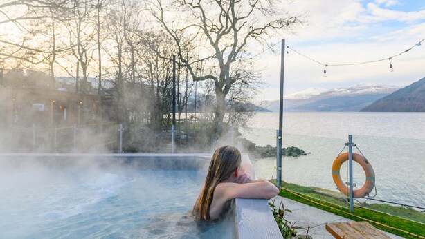 A person relaxing in a hot spring with a view of a lake and mountains in the background.