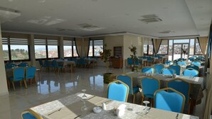 A spacious dining area with multiple tables and blue chairs, large windows offering a city view, and a polished floor. - Hasan Celebi Otel (Siverek)