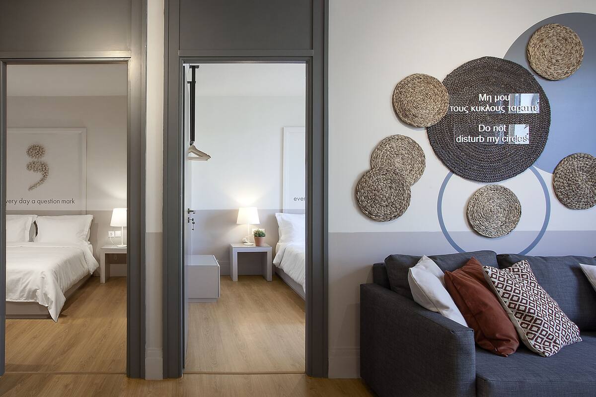 Une chambre d’hôtel moderne avec un lit, des tables de chevet, un canapé gris et des cercles décoratifs en fibres tressées accrochés au mur.