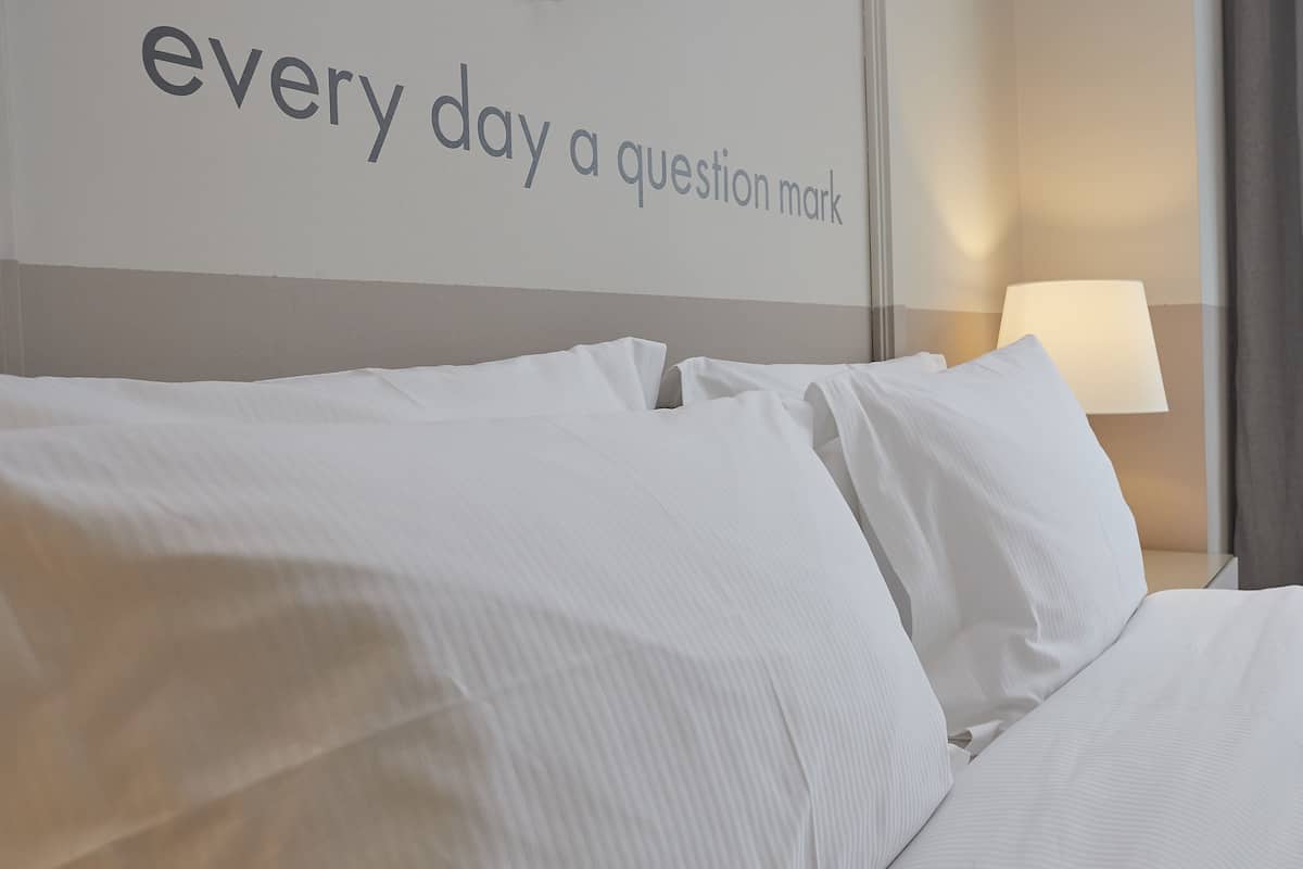 Une chambre d’hôtel avec un lit, des oreillers, un mur avec du texte, une lampe et une fenêtre avec des rideaux.