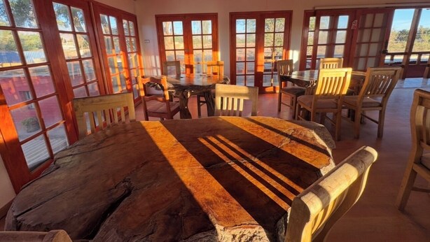 Een rustieke eethoek met een grote houten tafel en stoelen, grote ramen met houten kozijnen en uitzicht op de omgeving.