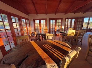 Un comedor rústico con una gran mesa de madera y sillas, ventanas amplias con marcos de madera, y vistas al exterior.