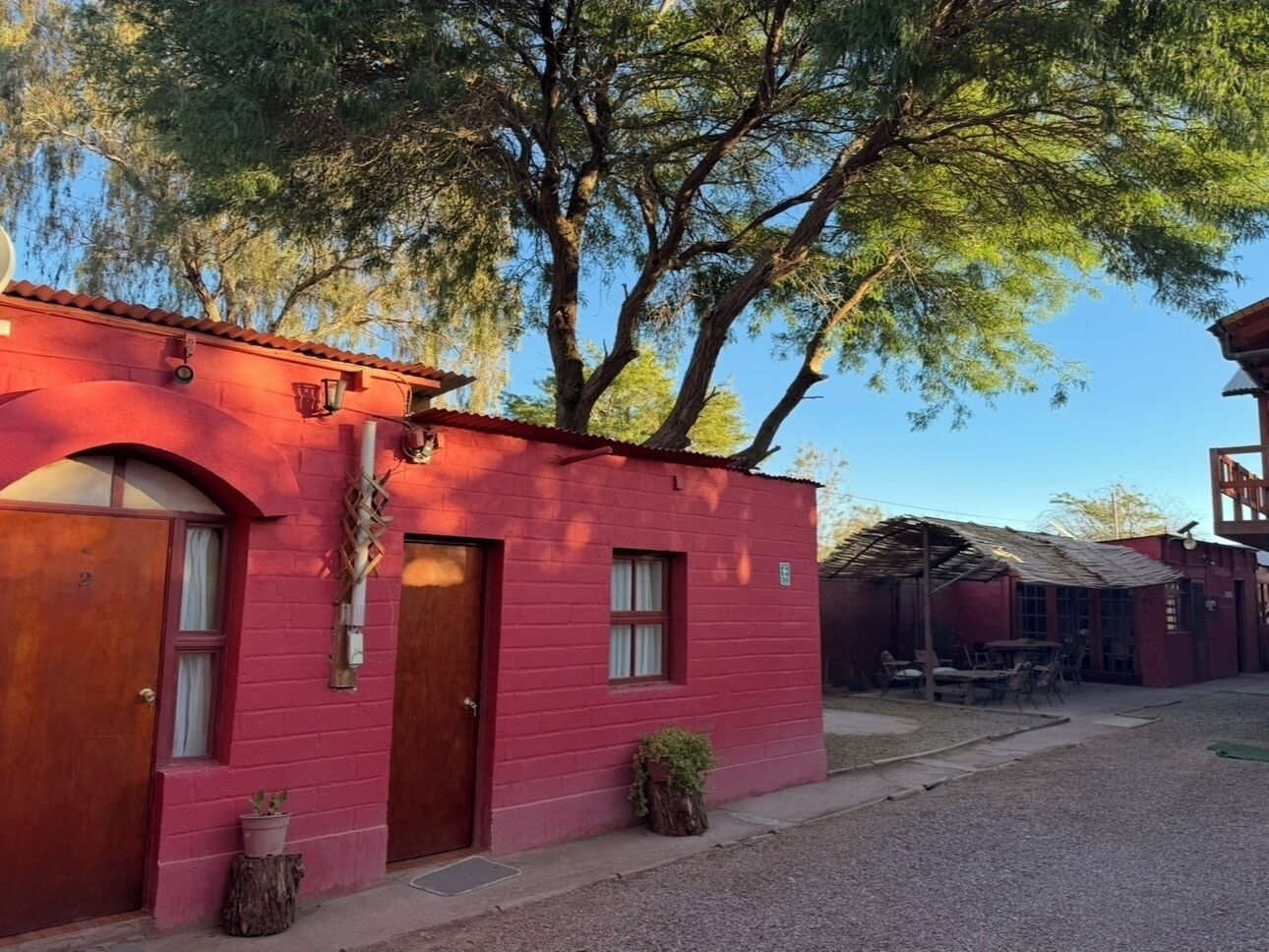 Un edificio rojo con dos puertas y ventanas, un pequeño porche con sillas y un árbol al fondo.
