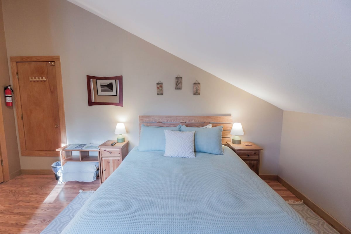 A bedroom with a sloped ceiling, a bed with a blue bedspread, two bedside tables with lamps, a wooden wardrobe, and a framed picture on the wall.