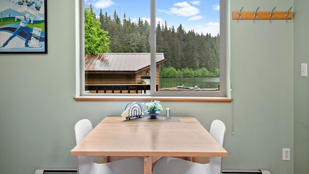 Ein Essbereich mit einem Holztisch und weißen Stühlen, ein Fenster mit Blick auf eine Hütte und einen Wald sowie ein an der Wand befestigter Garderobenständer.