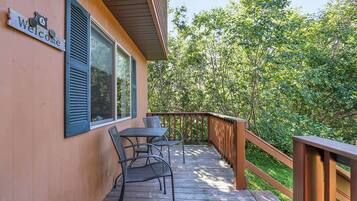 Une terrasse en bois avec une table et des chaises en métal, une pancarte « Bienvenue » et une vue sur une zone boisée.