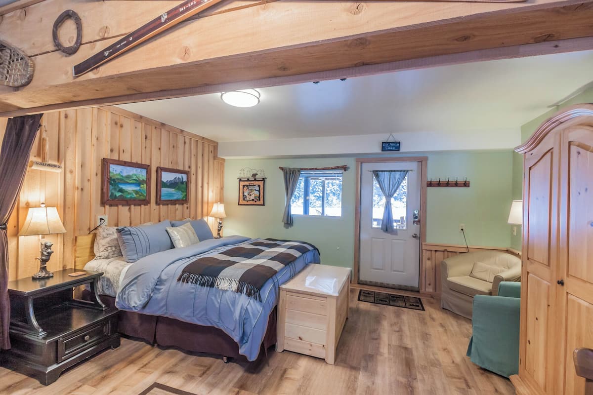 A cozy room with a wooden ceiling, a bed with a blue and white checkered bedspread, a wooden nightstand with a lamp, a green armchair, and a wooden wardrobe.