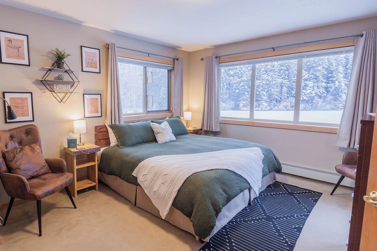A bedroom with a bed, two nightstands, a chair, a lamp, and a window with a view of snow-covered trees.