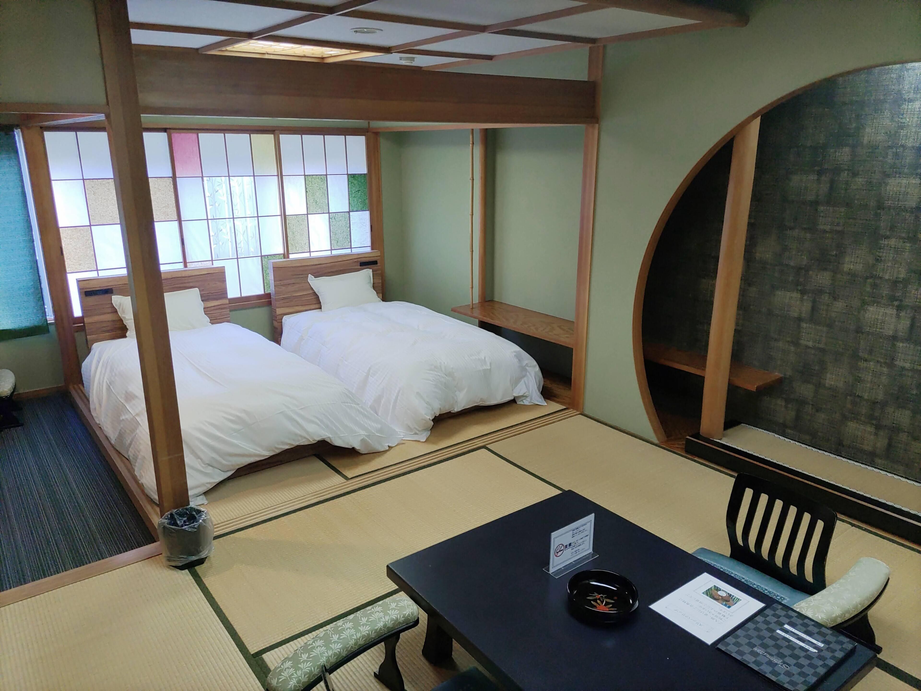 Ein traditioneller japanischer Raum mit Tatami-Boden, zwei Betten mit weißer Bettwäsche, einem kleinen Tisch mit einem schwarzen Tablett und Blick auf einen Garten durch einen Shoji-Bildschirm.