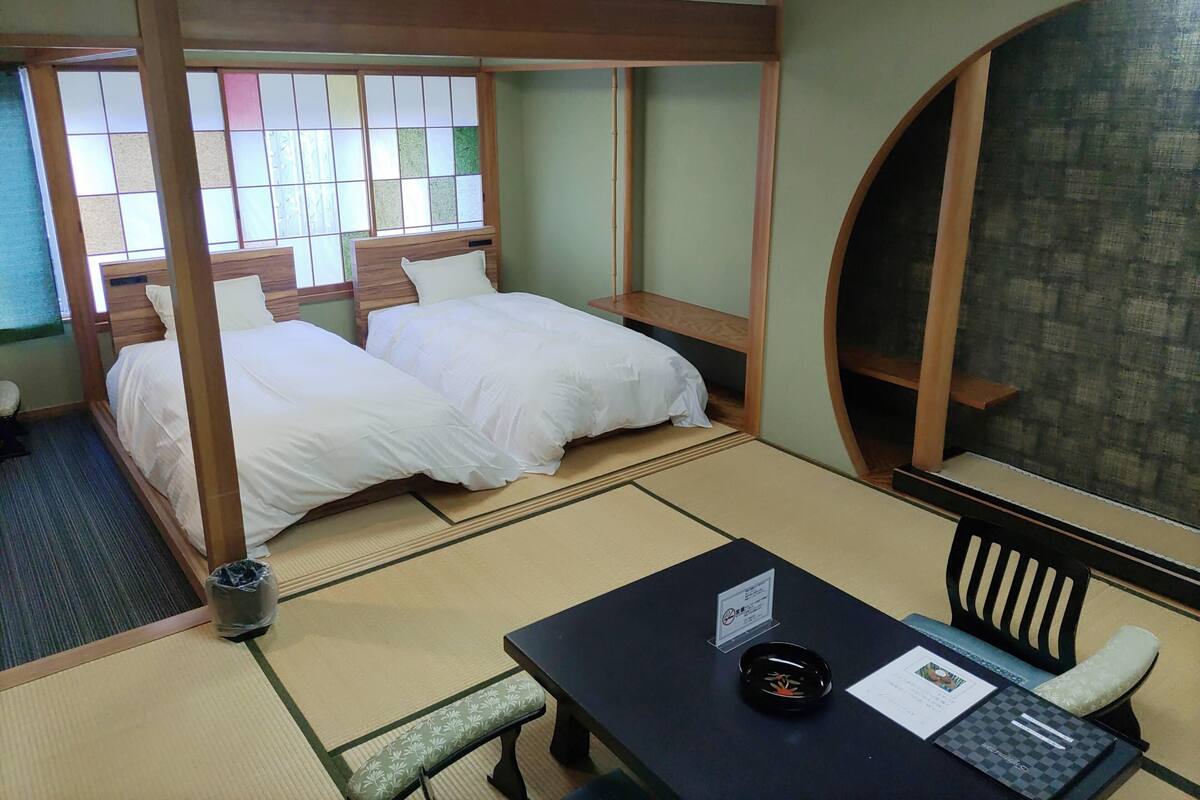 Een traditionele Japanse kamer met tatami vloer, twee bedden met witte beddengoed, een kleine tafel met een zwart dienblad, en een tuinzicht door een shoji scherm.