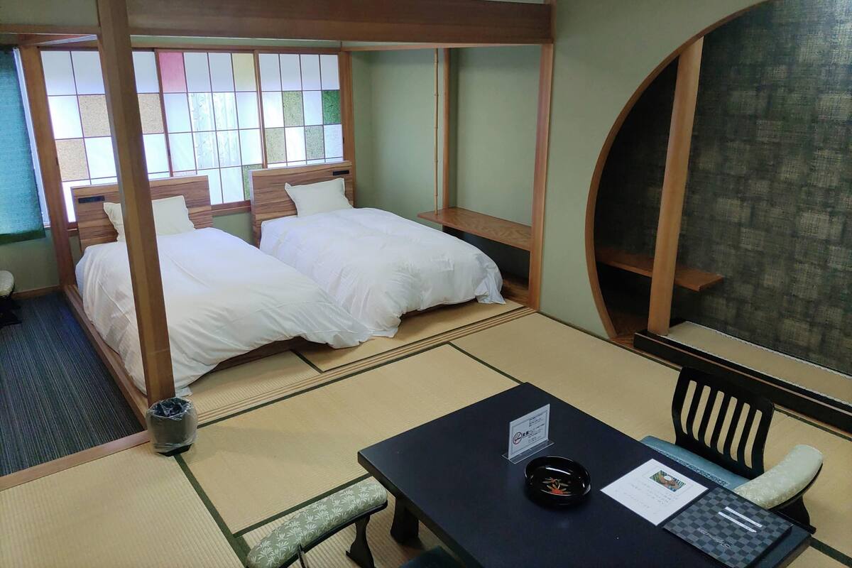 Een traditionele Japanse kamer met tatami vloer, twee bedden met witte beddengoed, een klein tafeltje met een zwarte dienblad, en uitzicht door een shoji scherm.