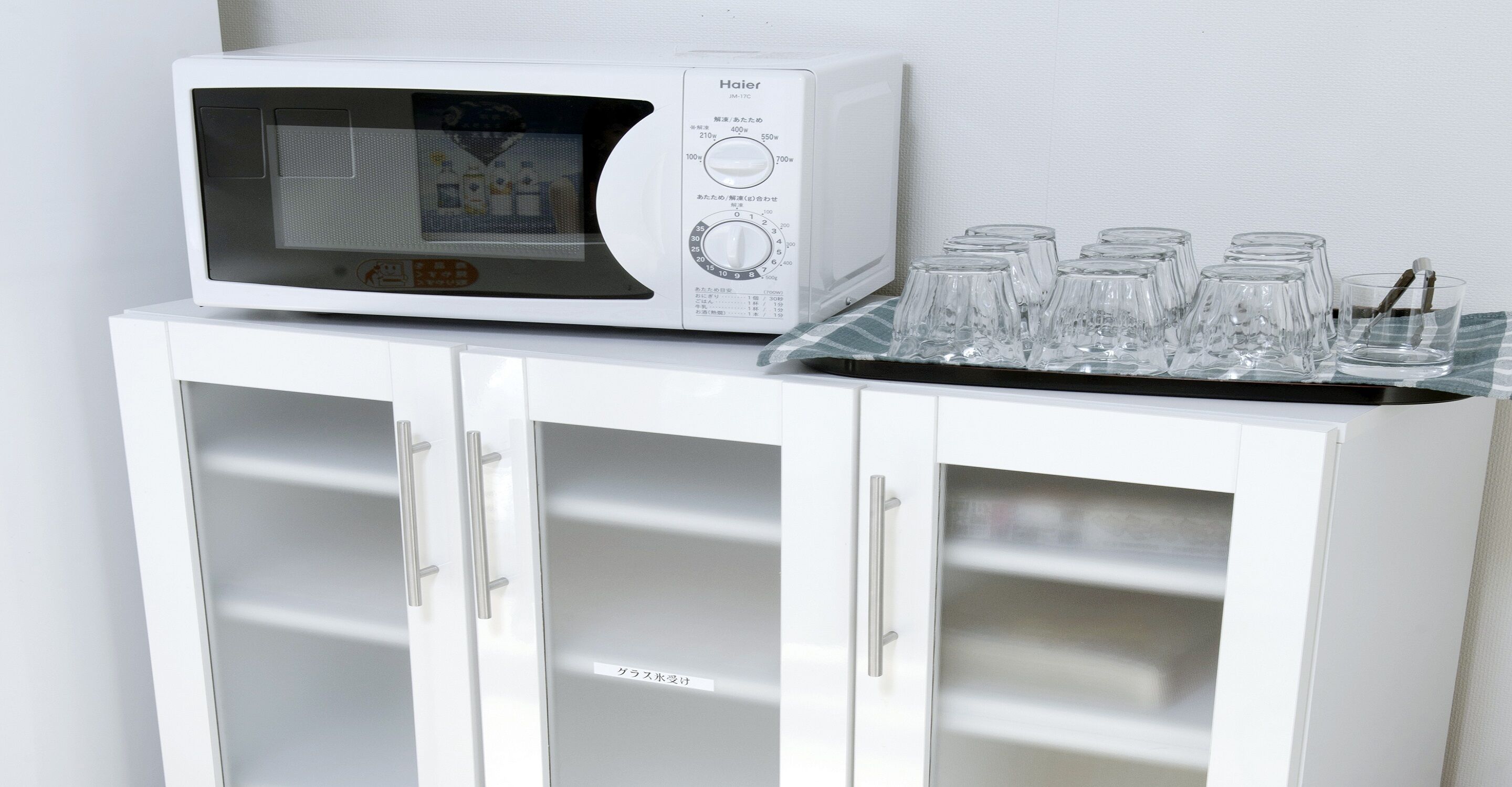 A white microwave on a cabinet with a tray of clear glasses.