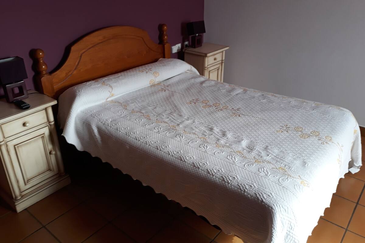 Un lit avec une tête de lit en bois, une couvre-lit blanche à motifs floraux et une table de chevet avec une lampe.