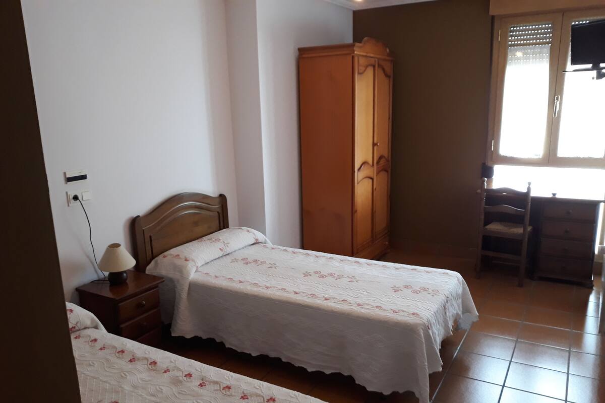 Une chambre avec deux lits simples, une armoire en bois, un bureau avec une chaise et une fenêtre avec des stores.