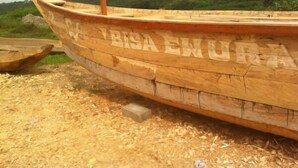 Un bateau en bois, portant l’inscription « VISA EWURIA » sur sa coque, est échoué sur une plage de sable, avec en arrière-plan une végétation luxuriante et une colline parsemée de bâtiments.