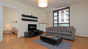 Ett modernt vardagsrum med en grå soffa, en platt-TV, ett soffbord och ett stort fönster med persienner.