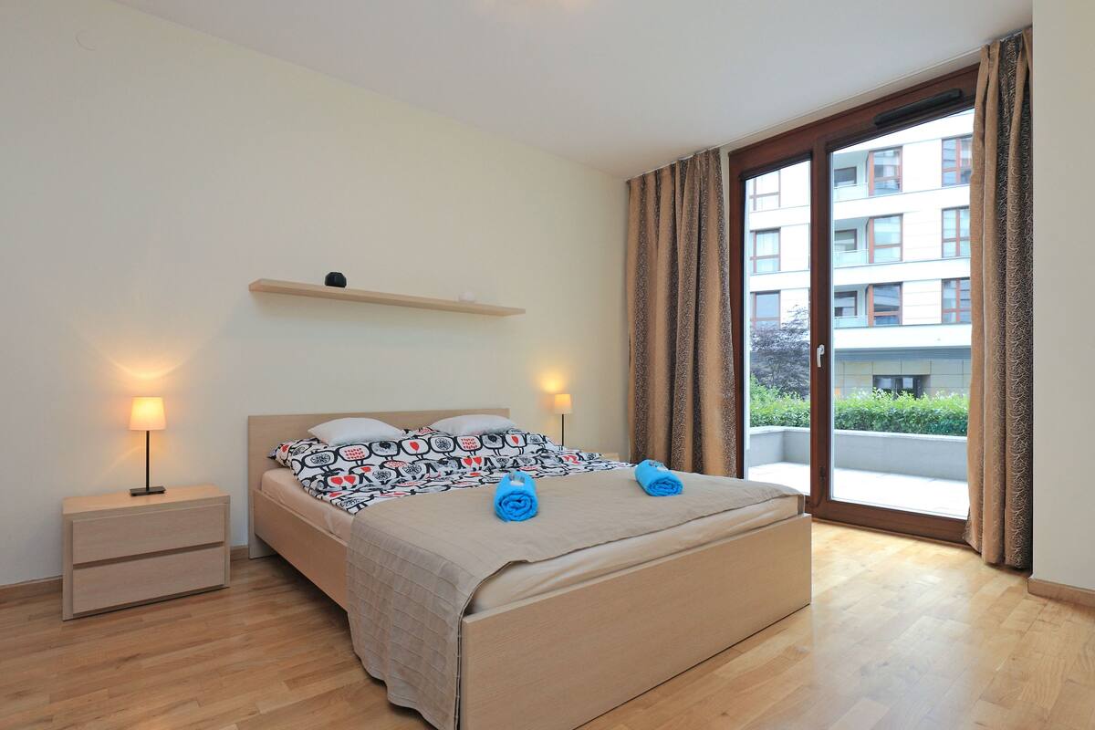A modern bedroom with a bed, bedside tables, a shelf, and a large window with curtains.
