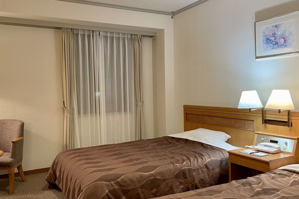 A hotel room with two beds, a chair, a desk with a lamp, and a window with curtains.