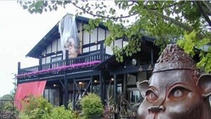 A building with a wooden facade, a balcony with purple flowers, and a garden with various plants.