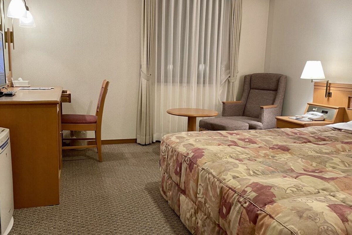Una habitación de hotel con una cama, un escritorio, una silla, una mesita y una mesita de noche con lámpara.