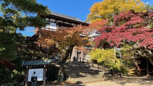 Un bâtiment traditionnel japonais, avec un escalier menant à l’entrée, entouré d’arbres aux couleurs automnales.