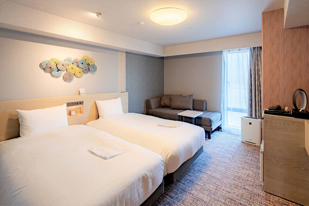 Habitación de hotel con dos camas, una zona de estar, una mesita pequeña y un ventanal grande con cortinas.